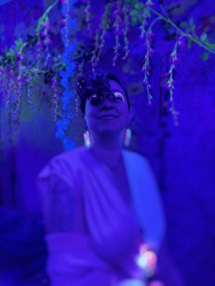 Beth wearing blacklight-glowing eyeshadow, earrings, and pendant, hair hanging over one eye, flowers hanging over and behind her.